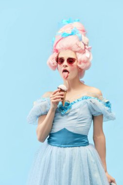 Genç prenses, güneş gözlüğü takan kraliçe dondurma yiyor ve mavi stüdyo arkaplanından kameraya bakıyor. Devir, modernlik ve rönesans kavramı, barok tarzı, güzellik, moda