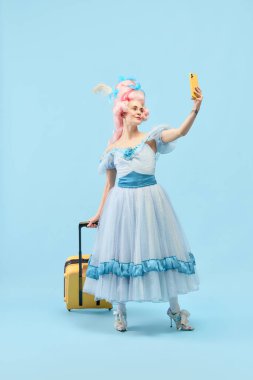 Gezgin blogcu. Mutlu büyüleyici prenses portresi, büyük pembe peruk takan kraliçe bavul tutuyor ve mavi stüdyo arka planında selfie çekiyor. Tatil kavramı, yaz, hafta sonu, internet, reklam.