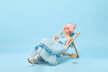 Yaz zamanı. Güzel prenses portresi, zarif elbiseler giyen kraliçe güneş yatağında oturuyor ve mavi arka planda güneşleniyor. Dönem karşılaştırması kavramı, rahatlatıcı, parti, eğlence, hafta sonu, reklam