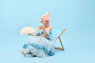 Sıcak yaz havası. Genç prensesin portresi, süslü elbiseler giyen güneş yatağında oturan ve mavi arka planda kokteyl içen kraliçe. Dönem karşılaştırması kavramı, rahatlatıcı, parti, seyahat, reklam