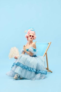 Sıcak yaz havası. Çekici prensesin portresi, süslü elbiseli kraliçe güneşli yatakta oturuyor ve mavi arka planda el yelpaze kullanıyor. Dönem karşılaştırması kavramı, rahatlatıcı, parti, seyahat, reklam