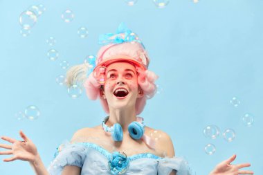 Portrait with princess, queen wearing dress playing with soap bubbles and smiling over blue studio background. Concept of comparison of eras, modernity, baroque style, beauty, fashion, human emotion clipart