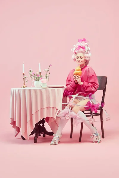 stock image Portrait with dreaming princess, queen wearing big wig and starting eat huge croissant on pink background with astonished face. Concept of food, diet, comparison of eras, modernity and renaissance