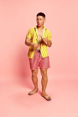 Çekici bir gencin portresi, öğrenci, plaj kıyafetleri ve güneş gözlüğü takan bir adam pembe renkli stüdyo arka planında asık suratlı. İnsan duyguları kavramı, ilişki, tatil, yaz, parti, reklam