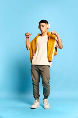 Genç ve çekici bir adamla portre, günlük tarz kıyafetler giyen, mavi arka planda kulaklıkla müzik dinleyen bir adam. Hobi kavramı, müzik, yaşam tarzı, moda, duygular, ad