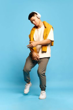 Genç ve çekici bir adamla portre, günlük tarz kıyafetler giyen, mavi arka planda kulaklıkla müzik dinleyen bir adam. Hobi kavramı, müzik, yaşam tarzı, moda, duygular, ad