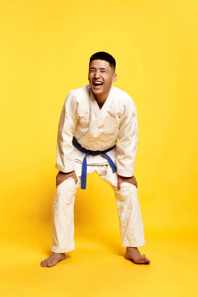 Portrait with cheerful young man, taekwondo, karate athlete wearing white kimono smiling over yellow color studio background. Concept of sport, education, skills, martial arts, healthy lifestyle