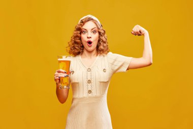 Zıplamanın gücü. Kaliteli bir elbise giyen, elinde bira bardağı tutan ve sarı arka planda gücünü gösteren genç bir kadın. İçki, alkol, degulasyon, festival, dinlenme, tatil, parti, reklam