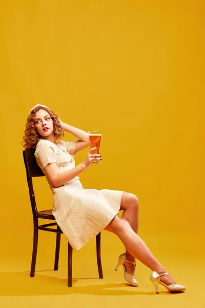 Chilled beer. One beautiful girl wearing stylish retro clothes drinking beer to cool off the heat over yellow background. Concept of drinks, alcohol, Oktoberfest, festivals, party, celebrating, ad