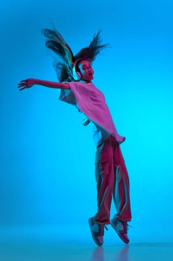 Etkileyici bir davranış. Sıradan kıyafetler ve kulaklıklar takan genç bir kızın portresi mavi arka planda neon ışıkta neşeyle müzik dinliyor. Güzellik kavramı, gençlik, insani duygular, rüyalar, reklam