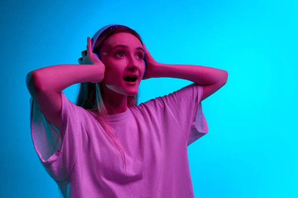 stock image Close up portrait of young girl, teenager in headphones and listening to music with open mouth over blue studio background in neon light. Concept of beauty, youth, human emotions, dreams, hobby, ad