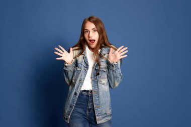 Şaşırmış biri. Pantolonlu, mavi renkli stüdyo arka planında ağzı açık bir şekilde el ele tutuşan hayrete düşmüş genç bir kızın portresi. İnsan duyguları kavramı, davranış, duygu çeşitliliği, reklam