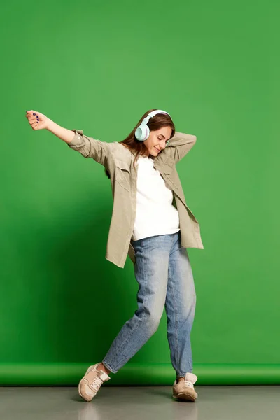 Stock image Music lover. Portrait of young girl, teenager wearing wireless headphones dancing and listening to music with joy over green background. Concept of beauty, youth, human emotions, dreams, hobby, ad