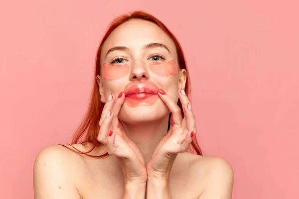 ピンクのスタジオの背景に唇のパッチで唇を潤す裸の肩を持つ若い赤毛の女の子と肖像画 美しさの概念 自然化粧 スキンケア 健康的な外観 化粧品 — ストック写真