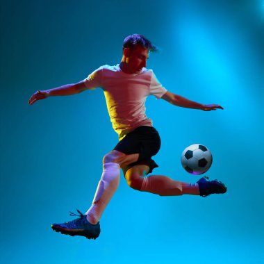 Hareket halinde. Futbolun dinamik portresi, spor üniformalı futbolcu neon ışıkta topu mavi arka planda tekmeliyor. Takım oyunu konsepti, spor, şampiyonluk, yarışma, zafer, reklam