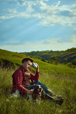 Sıcak yaz günü. Antika elbiseli bir adamın portresi çimenlerin üzerinde köpeğiyle oturan ve doğa manzarasında avlandıktan sonra dinlenen İngiliz spanyel köpeğiyle birlikte. Bahar kavramı, av, hobi, ad