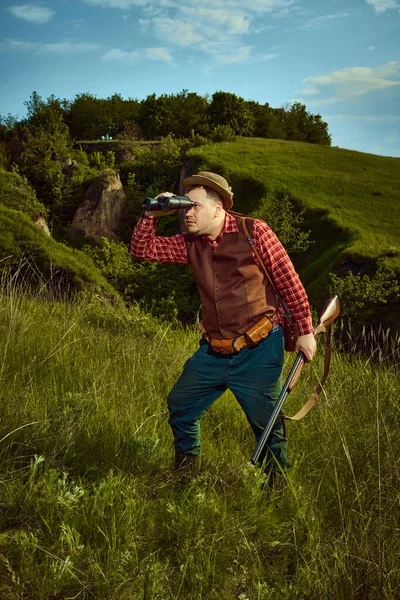 stock image Handsome man, hunter in hat with feather holding weapon and binoculars standing and watching, observing for prey over nature landscape background. Concept of hunting season, spring, prey, ad