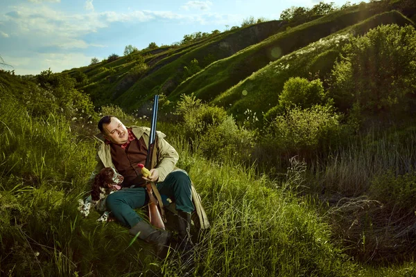 stock image Male hunter wearing vintage coat sitting with dog and holding prey over picturesque landscapes. Companion dog English springer spaniel. Concept of hunting season, prey, spring, hobby, ad