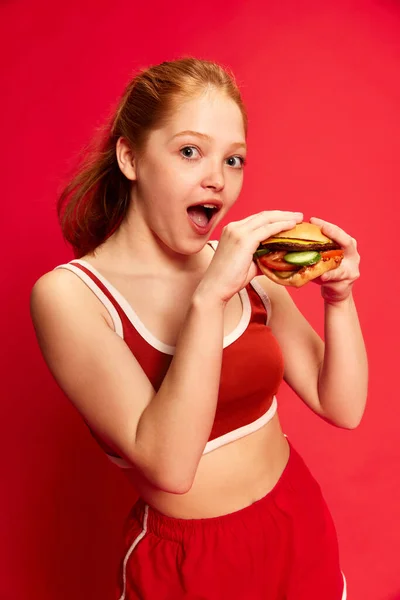 女孩穿着短裤 头戴着多汁的汉堡包 满脸喜色 略带红色的工作室背景 可用于食品广告 靠近点食物 广告的概念 — 图库照片