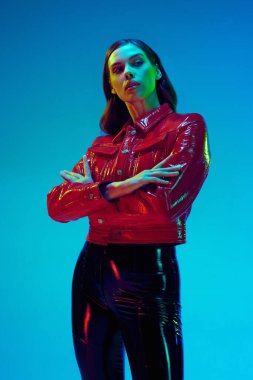 Çekici bir kadın, modaya uygun kıyafetler giyen, deri siyah pantolon giyen, mavi neon arka planda katlanmış elleriyle poz veren genç bir model. Güzellik kavramı, moda haftası, duygular, eğilimler, reklam