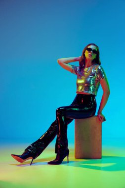 Yüksek model, modaya uygun kıyafetler giyen, deri pantolon giyen, parlak ışık altında mavi arka planda poz veren genç bir kadın. Güzellik kavramı, moda haftası, duygular, eğilimler, reklam