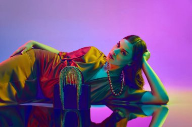 Güzel seksi kadın, model mor neon arka planda yerde yatıyor. Nazik kırmızı ipek elbise giyen, lüks bir çanta ve aksesuar kolye takan bir kız. Güzellik kavramı, duygular, reklam