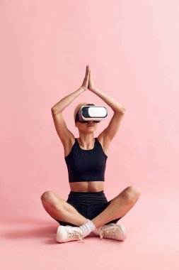 İnce vücutlu, siyah spor kıyafetleri ve VR gözlükleri olan genç bir kadın pembe stüdyo arka planında oturup meditasyon yapıyor. İnternetteki yoga. Spor yaşam tarzı, vücut ve sağlık, moda ve güzellik kavramı