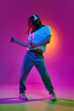 Günlük giysiler ve kulaklıklar takan genç bir kızın portresi neon ışıklarıyla pembe arka planda neşeli bir şekilde müzik dinliyor ve şarkının ritmiyle hareket ediyor. Gençlik kavramı, insani duygular, rüyalar, reklam