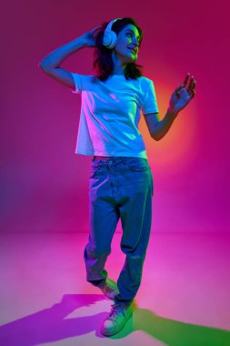 Hayalci kız. Mutlu, kısa saçlı, günlük kıyafetler giyen ve kulaklık takan, pembe arka planda neon ışıkta poz veren genç kadın portresi. Güzellik kavramı, gençlik, insani duygular, rüyalar, hobi