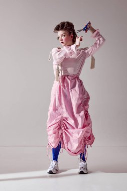 Değişiklikler. Pembe etekli, saçları makasla kesen güzel bir kızın portresi gri stüdyo arka planında. Kadın güzelliği, sanat, kadınlık, moda, duygular, tuhaflık kavramı