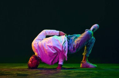 Esneklik. Spor kıyafetleri içinde hip-hop dansı yapan genç bir adam, neon ışıkta siyah stüdyonun arka planına karşı breakdance. Sokak dansı konsepti, moda, gençlik, hobi, dinamik, reklam