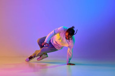 Dehşet verici kıyafetleri olan, hip-hop dansı yapan, neon ışıkta rengarenk stüdyo arkaplanına karşı breakdance yapan genç bir adam. Sokak dansı konsepti, moda, gençlik, hobi, dinamik, reklam