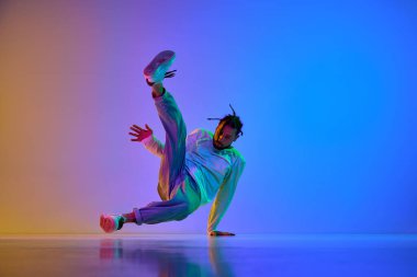 Dehşet verici kıyafetleri olan, hip-hop dansı yapan, neon ışıkta rengarenk stüdyo arkaplanına karşı breakdance yapan genç bir adam. Sokak dansı konsepti, moda, gençlik, hobi, dinamik, reklam