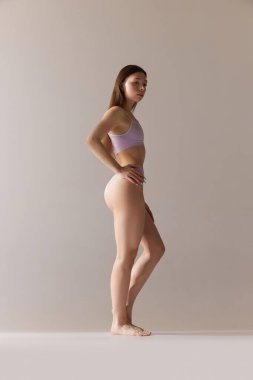 Gri stüdyo arka planında iç çamaşırlarıyla poz veren zayıf bir kızın tam boy portresi. Vücut şekline özen gösteriyorum. Doğal güzellik kavramı, cilt bakımı, sağlık, sağlık, kadınlık. Ad