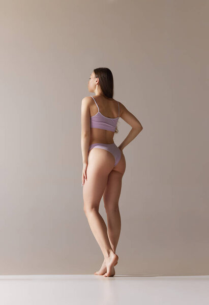 Full-length back view portrait of young girl with slim body, buttocks posing in underwear over grey studio background. Concept of natural beauty, body and skin care, health, wellness, femininity. Ad