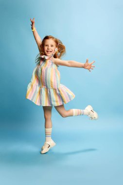 Kızıl saçlı güzel bir kızın tam boy portresi, şirin çizgili elbiseli bir çocuk ve mavi stüdyo arka planına neşeyle zıplıyor. Çocukluk kavramı, duygular, yaşam tarzı, moda, neşe. Ad