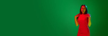 Şaşırt ve şaşırt. Neon ışıkta yeşil arka planda poz veren şık kırmızı elbiseli güzel bir genç kızın portresi. Gençlik kavramı, insani duygular, yaşam tarzı, moda. Pankart. İlan için alanı kopyala