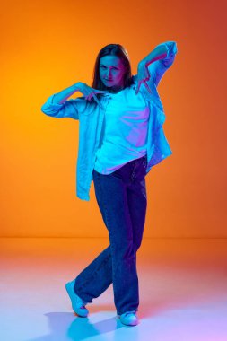 Neon ışıklarıyla gradyan turuncu stüdyo arkaplanına poz veren rahat giyimli genç güzel bir kadının tam boy portresi. Gençlik kavramı, insan duyguları, yaşam tarzı, yüz ifadesi, reklam