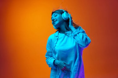 Kulaklıkla müzik dinleyen genç, gülümseyen bir kadın. Neon ışıklarıyla arka plandaki turuncu renkli stüdyoya karşı. Enfes. Gençlik kavramı, insan duyguları, yaşam tarzı, yüz ifadesi, reklam