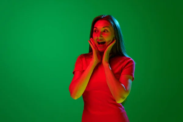 stock image Happy shocked face. Portrait of young beautiful woman in red dress posing against green background in neon light. Celebration. Concept of youth, human emotions, lifestyle, facial expression, ad