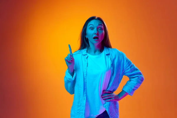 stock image Young emotional woman raising finger up meaning ideas and thoughts against gradient orange studio background in neon light. Concept of youth, human emotions, lifestyle, facial expression, ad