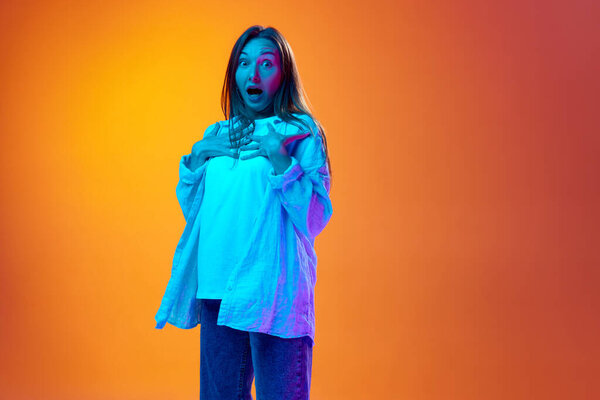 Shock and surprise. Portrait of beautiful young girl in casual clothes posing over gradient orange studio background in neon light. Concept of youth, human emotions, lifestyle, facial expression, ad