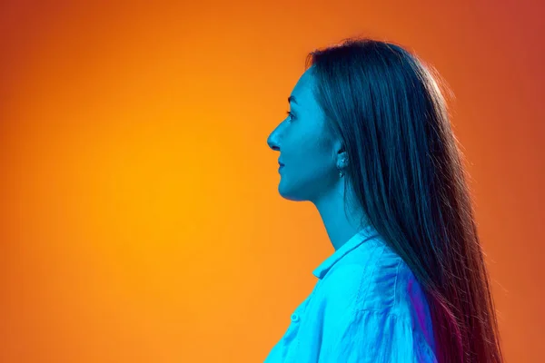 stock image Side view portrait of young woman in casual clothes posing against gradient orange studio background in neon light. Profile. Concept of youth, human emotions, lifestyle, facial expression, ad