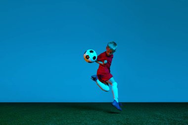 Spor üniformalı bir futbolcunun mavi arka planda futbol topuyla zıpladığı bir stüdyo fotoğrafı. Eylem kavramı, spor yaşam tarzı, takım oyunu, sağlık, enerji, canlılık ve reklam