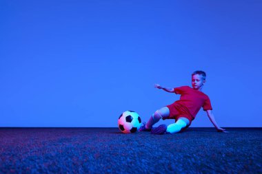 Sportif bir çocuk, kırmızı üniformalı bir çocuk neon ışıkta futbol antrenmanı yapıyor. Eylem kavramı, spor yaşam tarzı, takım oyunu, sağlık, enerji, canlılık ve reklam