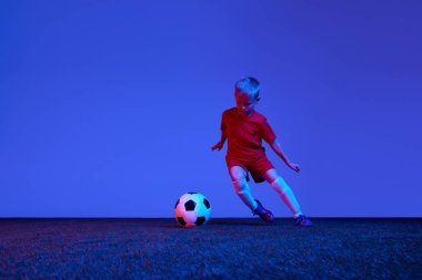 Sportif bir çocuk, kırmızı üniformalı bir çocuk neon ışıkta futbol antrenmanı yapıyor. Eylem kavramı, spor yaşam tarzı, takım oyunu, sağlık, enerji, canlılık ve reklam