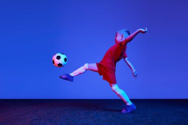 Sportif bir çocuk, kırmızı üniformalı bir çocuk neon ışıkta futbol antrenmanı yapıyor. Eylem kavramı, spor yaşam tarzı, takım oyunu, sağlık, enerji, canlılık ve reklam