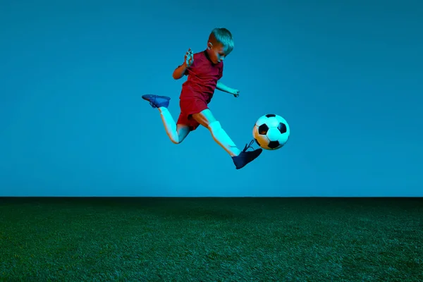 演播室拍摄的小男孩 身穿运动服的足球运动员在霓虹灯下跳过蓝色背景的足球 行动概念 运动生活方式 团队精神 活力及广告 — 图库照片