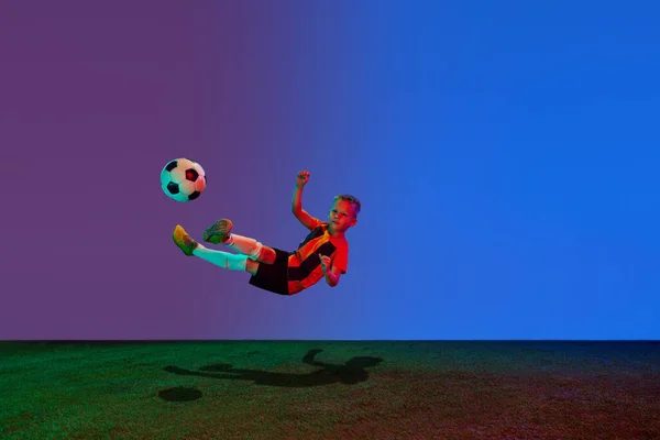 小さな男の子のスタジオショット ネオンで青の背景にサッカーボールと一緒にジャンプするスポーツユニフォームのサッカー選手 アクションの概念 スポーツライフスタイル チームゲーム エネルギー 活力と広告 — ストック写真