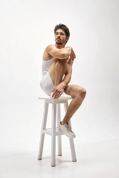 工作室拍摄年轻迷人的白人男子穿着家居服坐在白色背景的高椅子上 男性健康 男性美体 广告的概念 — 图库照片